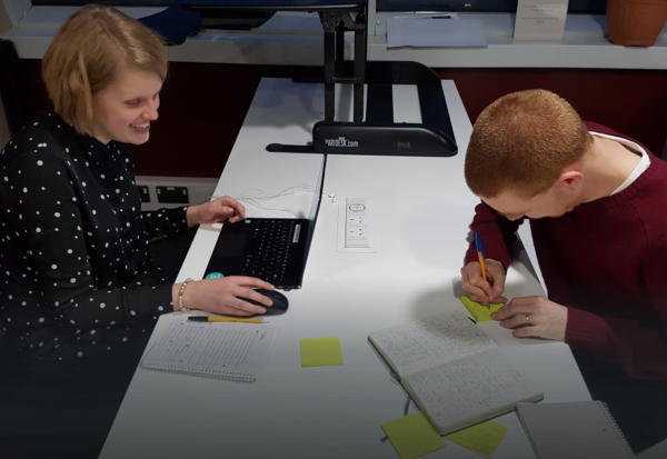 Two people opposite each other at a desk, one on a laptop the other writing a post-it note.