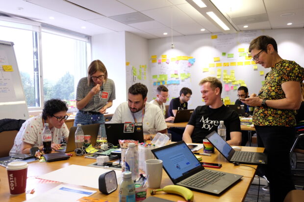A group of product managers and transformation leads take part in a design sprint. There are two tables with a team gathered around each with sticky notes and laptops.