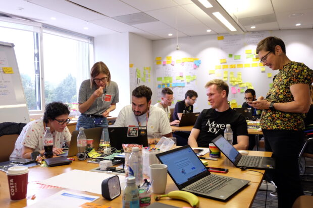 A group of product managers and transformation leads take part in a design sprint. There are two tables with a team gathered around each with sticky notes and laptops.